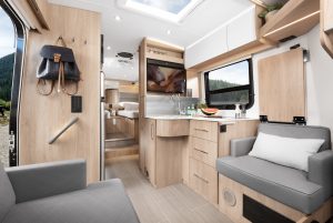 The living room area on the inside of a motorhome features grey seating and light-wood cabinets.