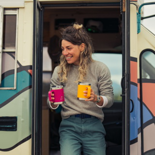 A woman exits a travel trailer, holding two Dometic Thermo Mugs